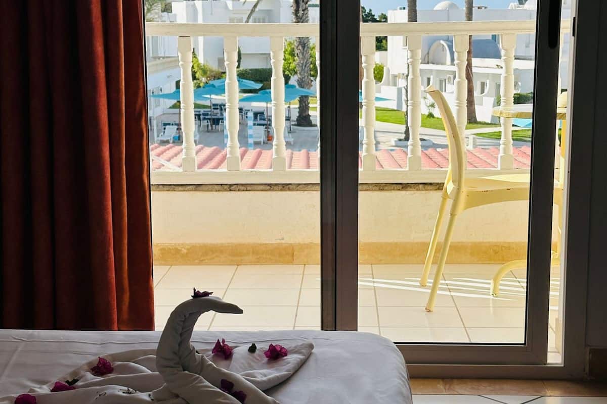 A hotel room with a bed, a balcony with a white railing, and a view of palm trees and a pool area.