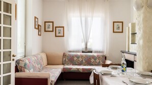 Uma sala de jantar com mesa posta para quatro pessoas, um sofá com um colcha colorida e uma janela com cortinas leves.