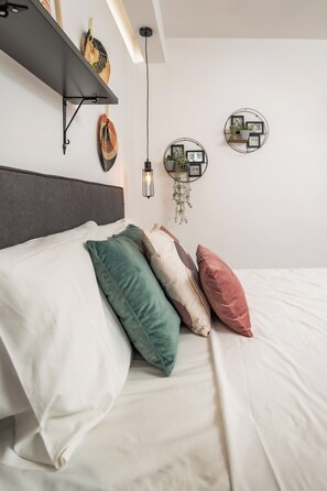 Ein ordentlich bezogenes Bett mit weißen und farbigen Kissen, einem dunklen Kopfteil und an der Wand befestigten Regalen mit dekorativen Accessoires.