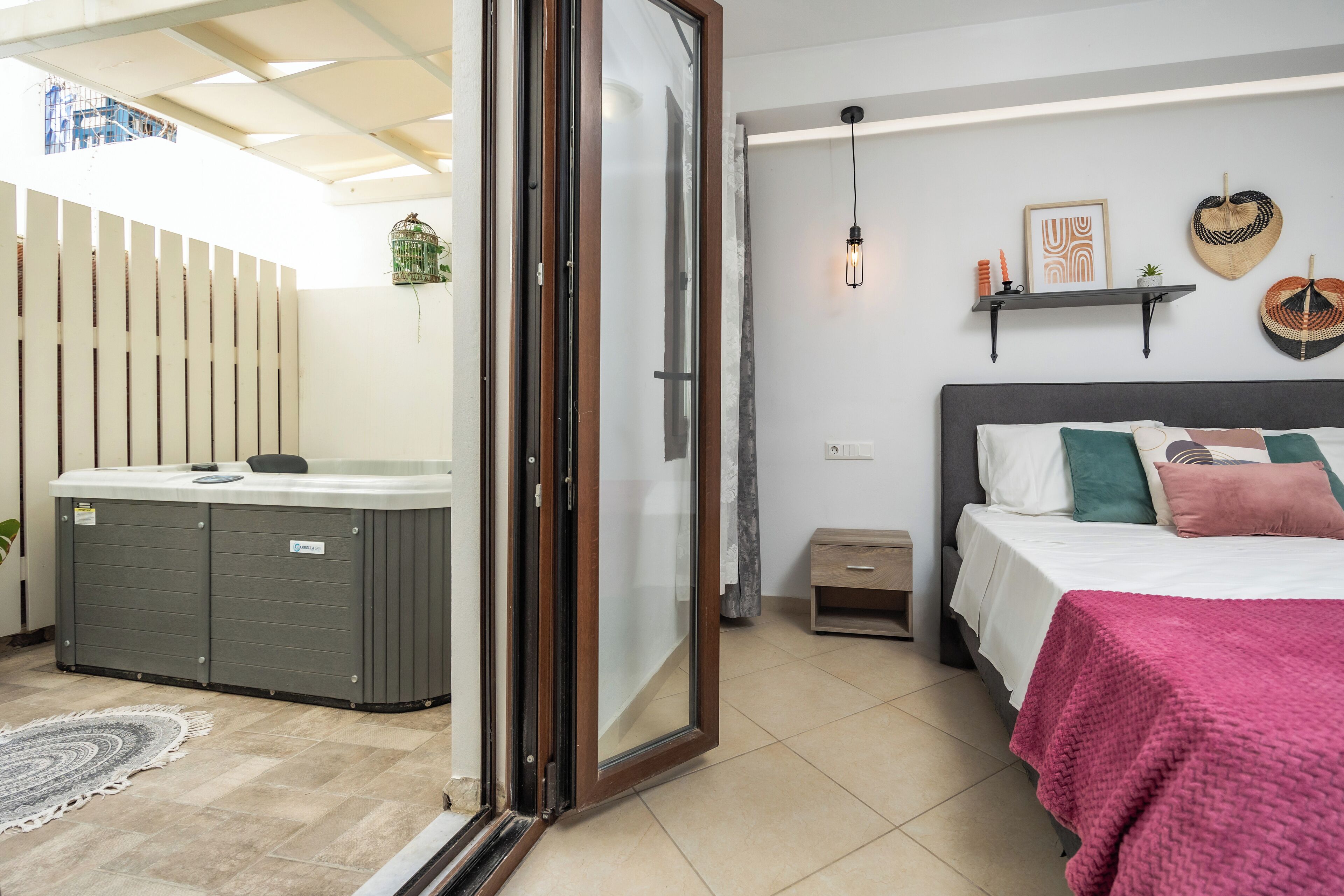 A bedroom with a bed, bedside table, and a small nightstand. There is a window with a view of the outside, a ceiling fan, and a wall-mounted shelf with decorative items.