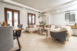 A modern living room with a sofa, armchair, dining table, and a small coffee table. There is a window with a view of the outside, and an air conditioning unit mounted on the wall. - Naxos Chora Modern Dream w Patio Jacuzzi (Naxos)