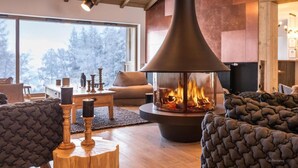Ein gemütliches Wohnzimmer mit großem Kamin, Holzmöbeln und Blick auf Bäume durch raumhohe Fenster.