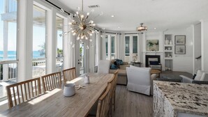 A spacious living room with a wooden dining table, comfortable seating, a fireplace, and large windows offering a view of the ocean. - Gulf-front, Private Pool ~ Stunning Gulf Views From All Levels + Beach Setup (Panama City Beach)