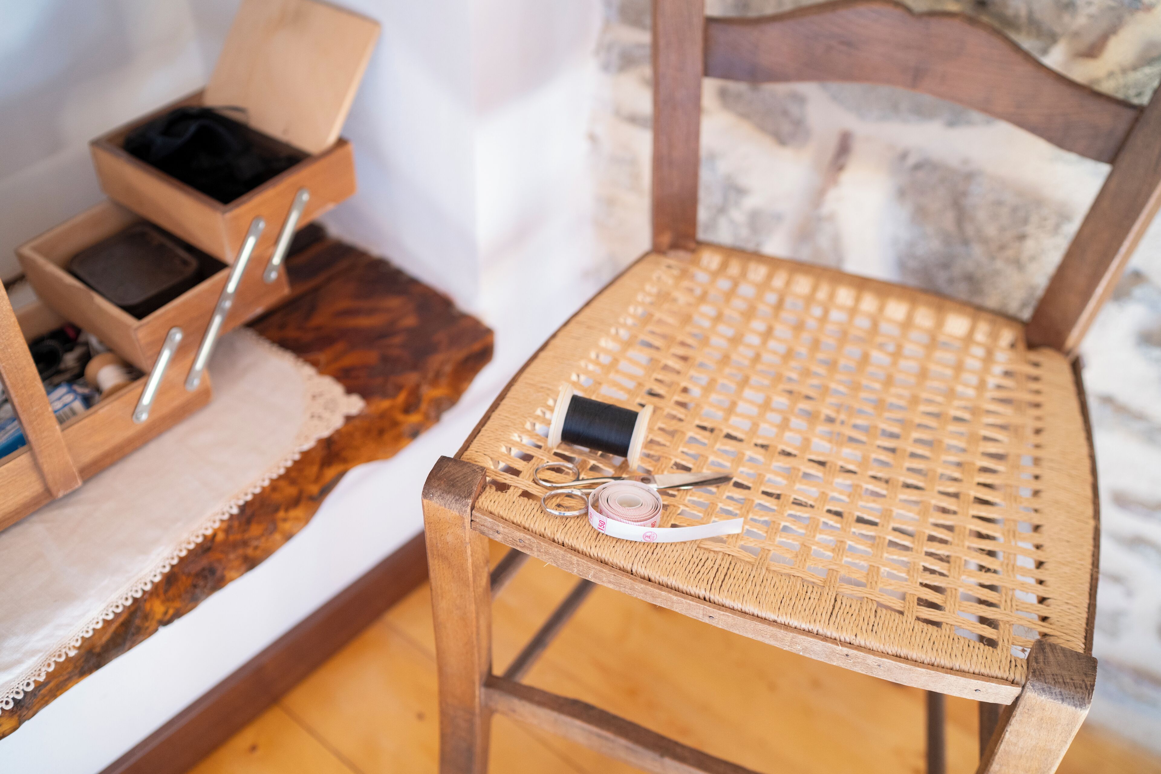 Uma cadeira de madeira com assento e encosto trançados, uma pequena caixa de madeira com divisórias e um novelo de linha no apoio de braço da cadeira.