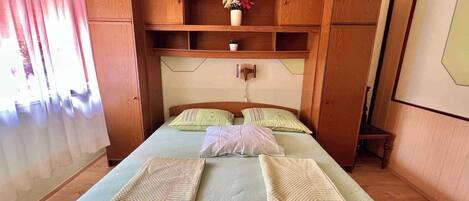 A bedroom with a bed, wooden wardrobe, and a small shelf with a vase.