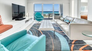A modern living room with a large TV, teal and white furniture, a glass coffee table, and a view of the ocean through large sliding doors. - Beach House 104A - Gulf Front Resort! (Miramar Beach)