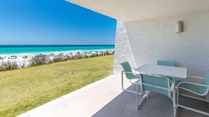 A patio with a white table and chairs, overlooking a beach with umbrellas and a clear blue sky. - Beach House 104A - Gulf Front Resort! (Miramar Beach)