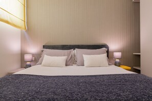 Ein ordentlich bezogenes Bett mit grauer Decke, zwei Nachttischlampen und einem Kopfteil mit dunkler Akzentfarbe.
