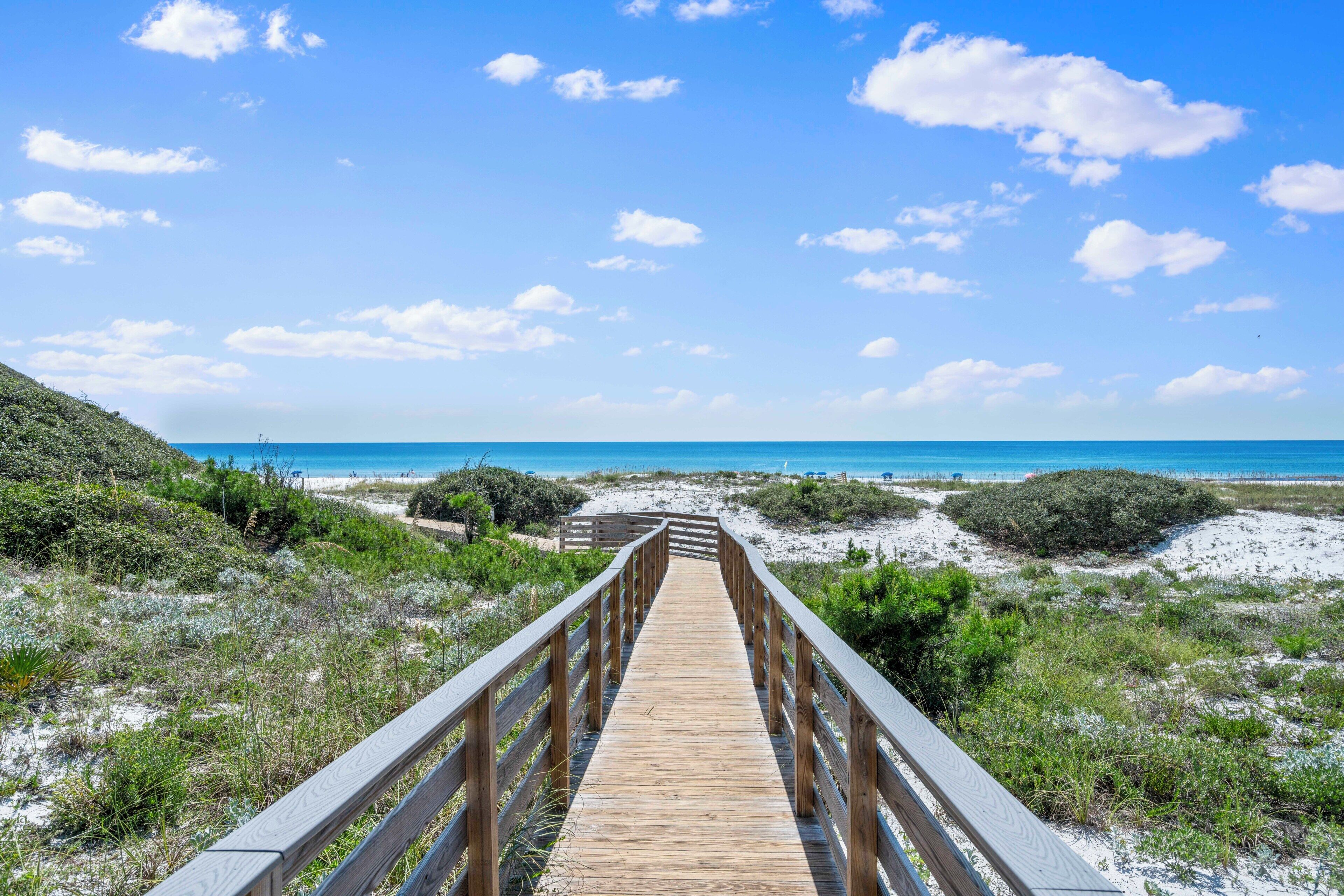Ein hölzerner Steg führt zu einem Strand mit blauem Himmel und spärlicher Vegetation.