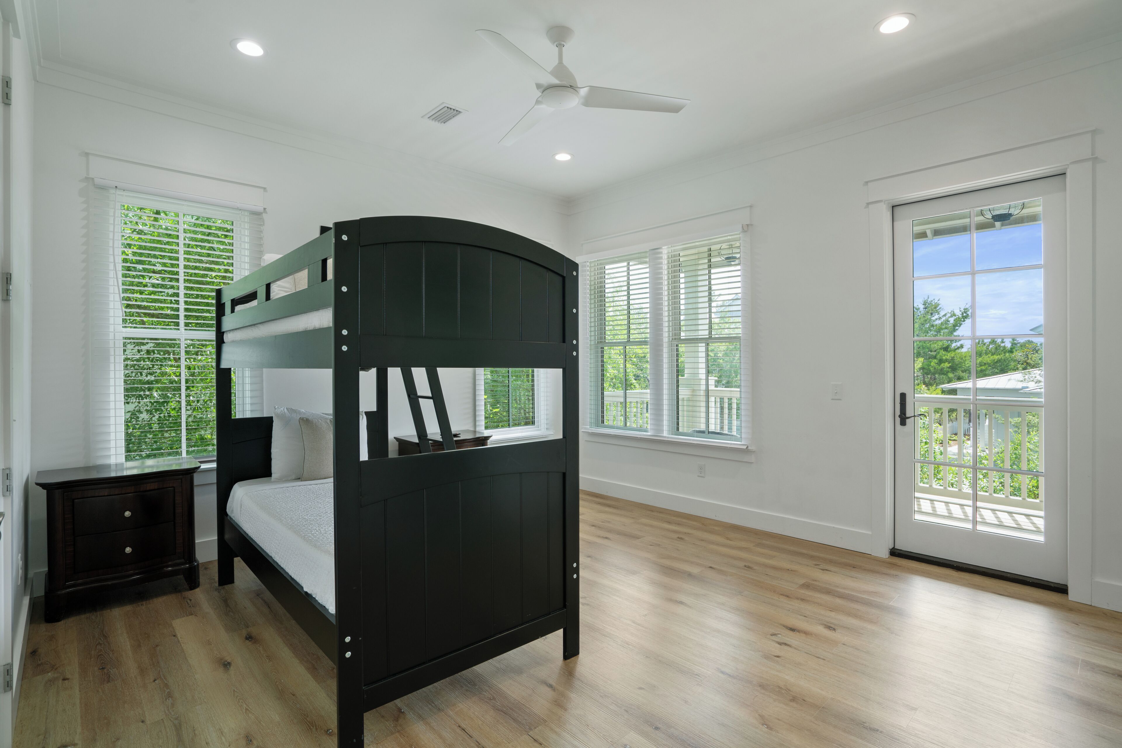 Une chambre avec un lit superposé, une table de chevet, un ventilateur de plafond et une vue sur l’extérieur, visible à travers une porte.