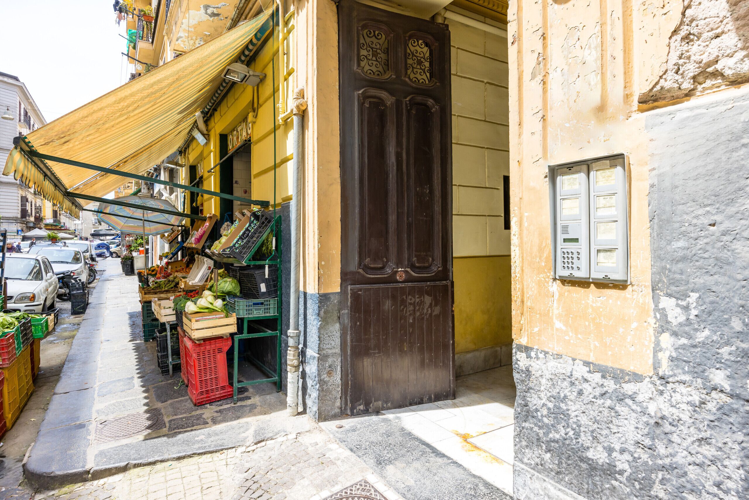 Una calle estrecha con un puesto de mercado que vende productos frescos, una tienda cerrada con una puerta de madera oscura y un cuadro eléctrico en la pared.
