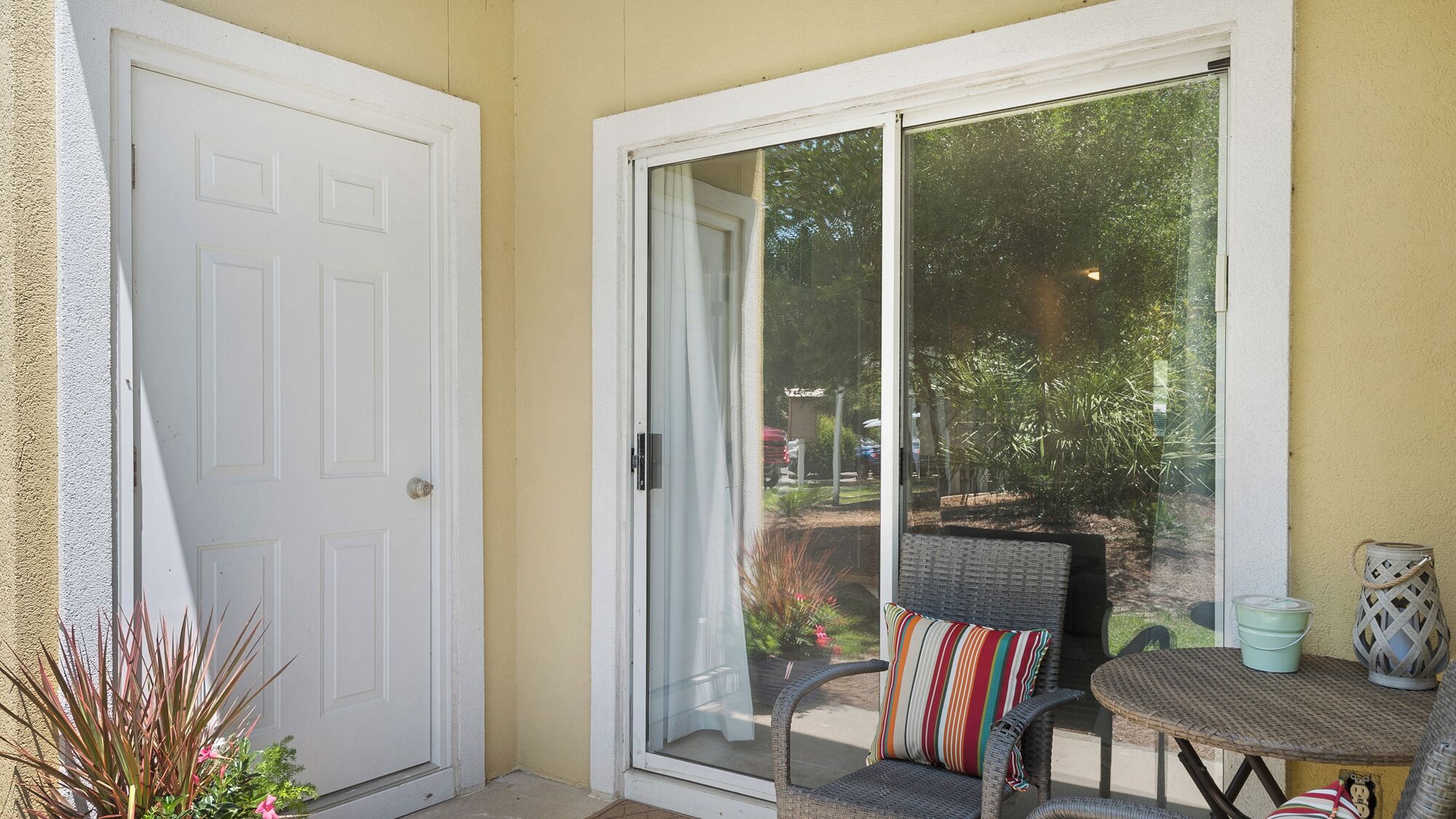 A room with a sliding glass door, a wicker chair with a striped cushion, a small round table, and a potted plant.