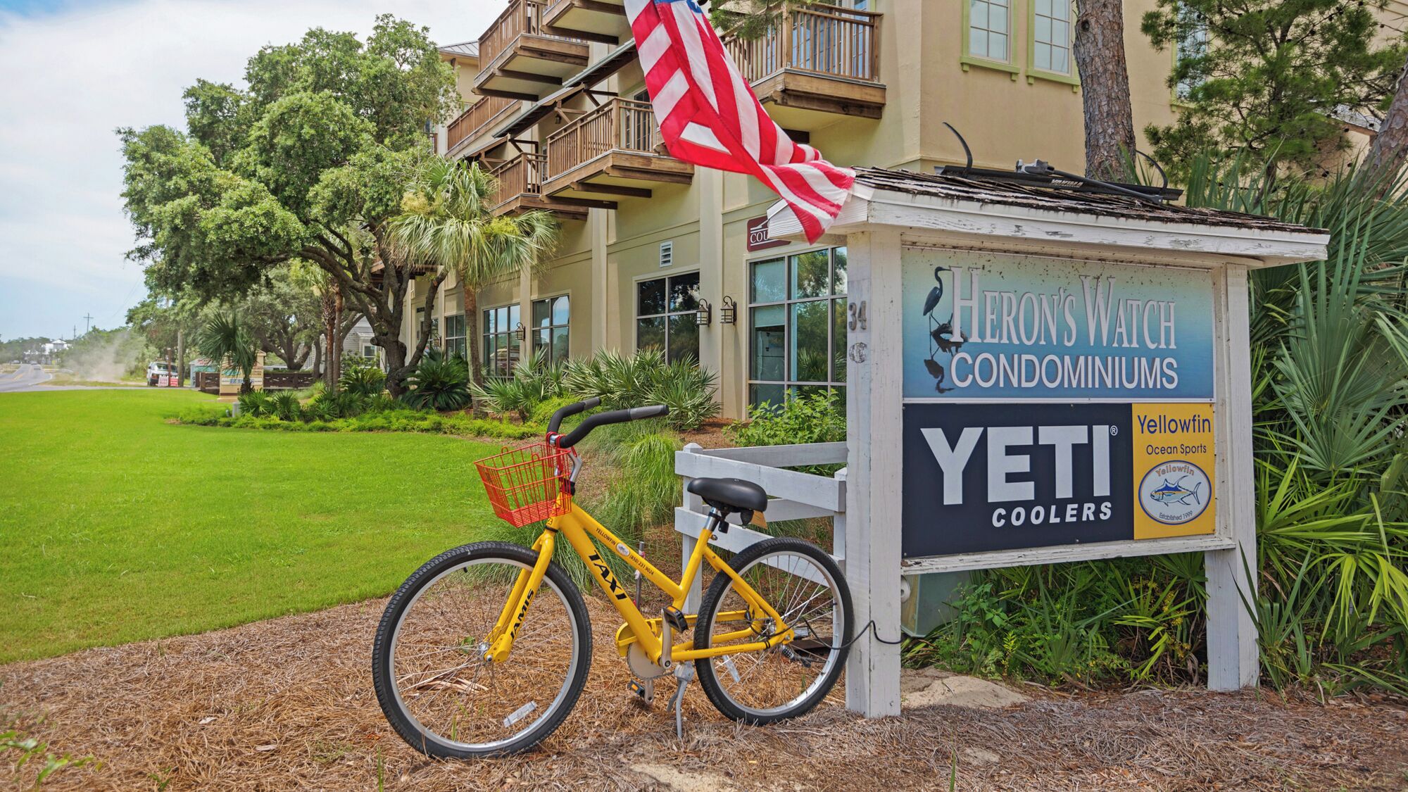 A yellow bicycle parked in front of a building with a sign advertising 'Heron's Watch Condominiums' and 'Yeti Coolers'.