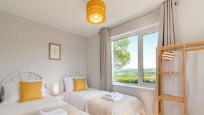 Une chambre avec deux lits, une table de chevet avec une lampe, une étagère en bois en forme d’échelle et une fenêtre donnant sur un paysage verdoyant.