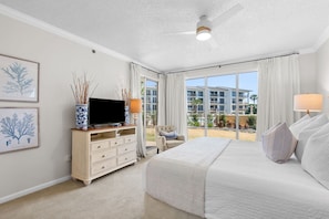 A hotel room with a large bed, a TV on a wooden cabinet, a chair, and a balcony with a view of buildings. - 1st Floor Gulf Side- Lagoon Pool- Beach Chair Setup (Panama City Beach)