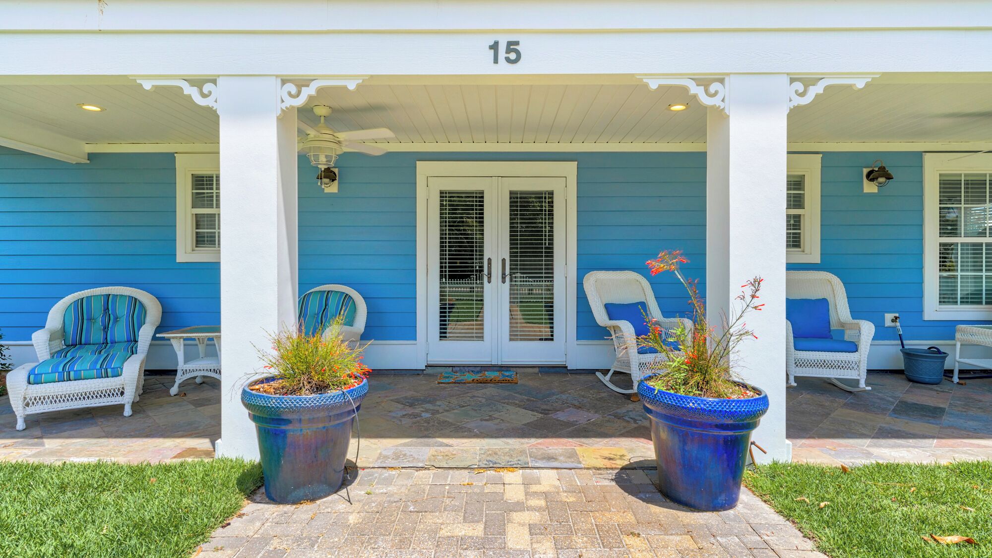 A blue house with a porch, numbered 15, featuring two white wicker chairs with blue cushions, a white door, and potted plants.