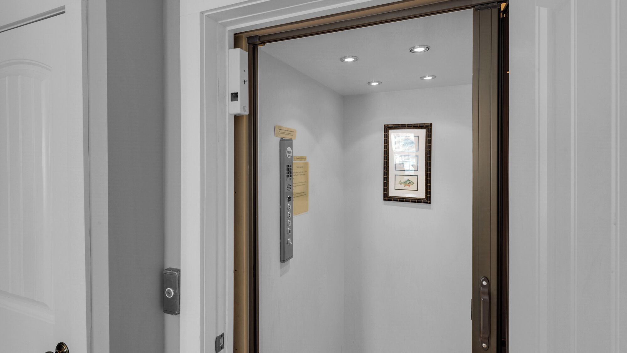 A hotel room with a wooden door, a wall-mounted thermostat, a framed picture, and a small window.