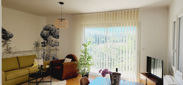 Een woonkamer met een bank, fauteuil, salontafel, televisie en groot raam met zonwering.