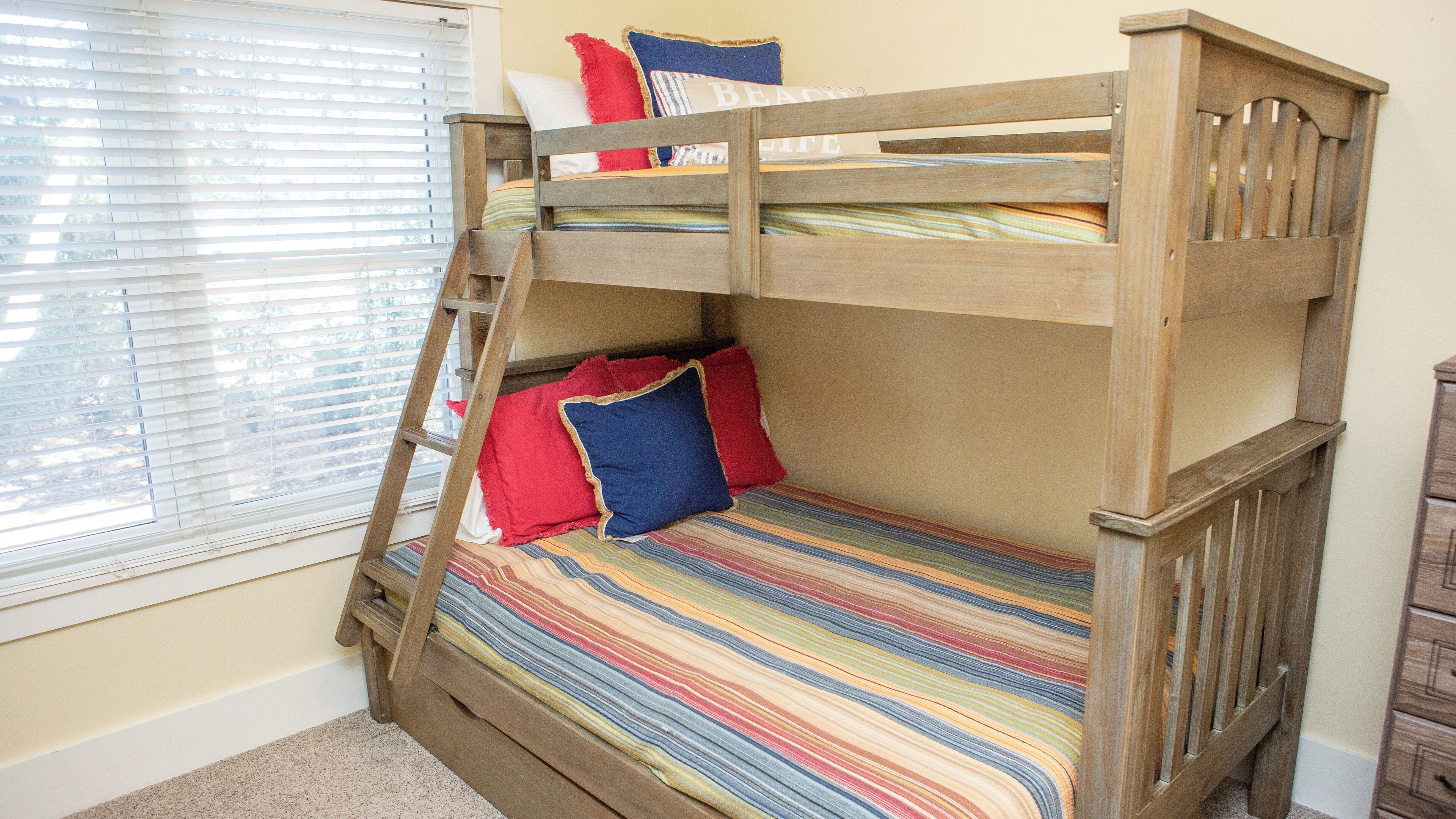A bunk bed with a striped bedspread and pillows, a wooden ladder, and a window with blinds.