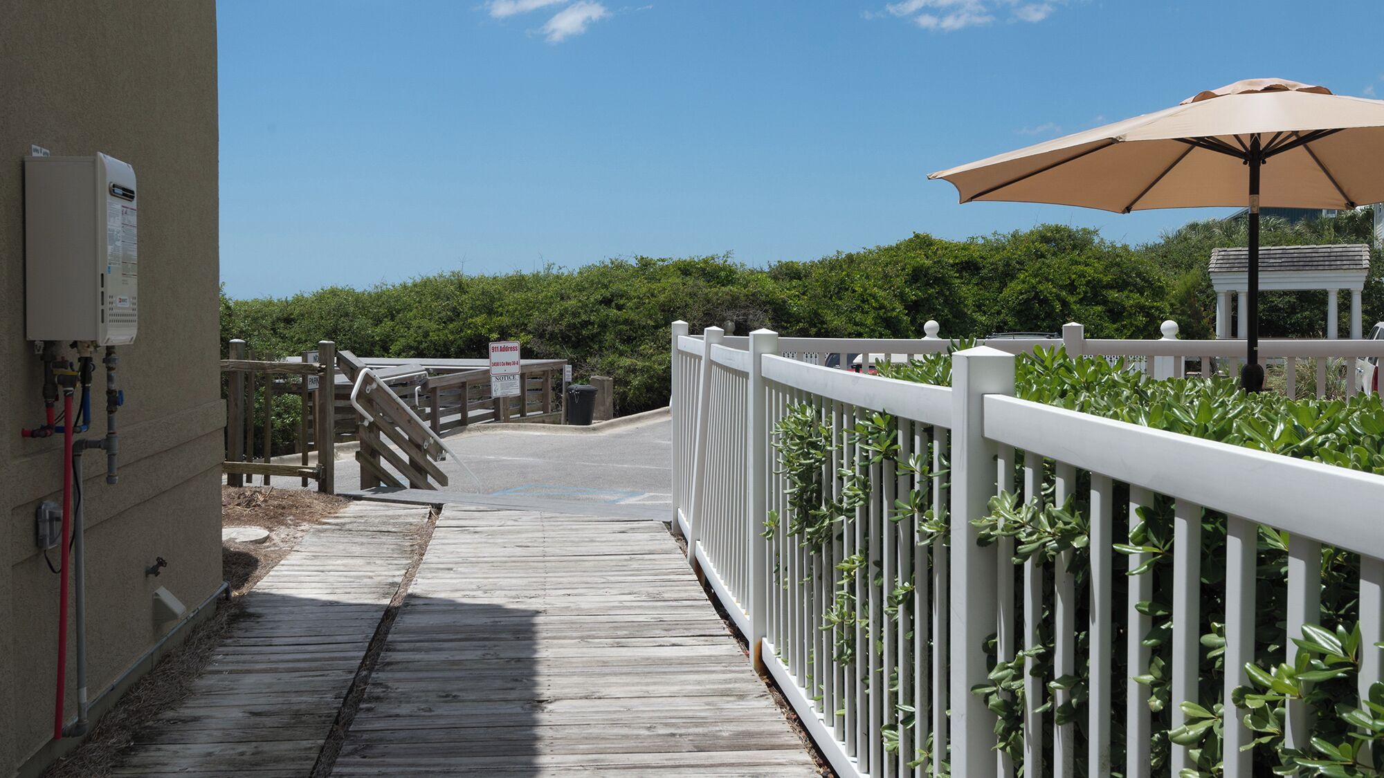 A wooden pathway leading to a gated entrance with a white fence and greenery on the right.