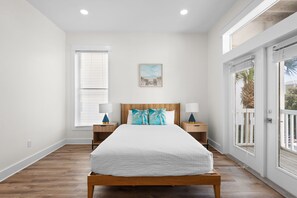 Quarto com cama de madeira, roupa de cama branca, duas mesas de cabeceira com abajures, uma janela com persianas e uma porta para área externa.