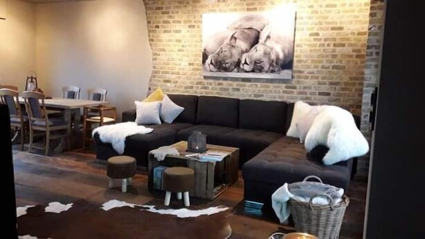 A living room with a dark-colored sofa, a dining table with chairs, a brick wall, and a painting of animals.