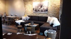 A living room with a dark-colored sofa, a dining table with chairs, a brick wall, and a painting of animals. - Belle Vie Guesthouse near Bruges (Oostkamp)