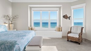 Une chambre avec un grand lit, un fauteuil en osier, une vue sur l’océan et un vase rempli de fleurs.