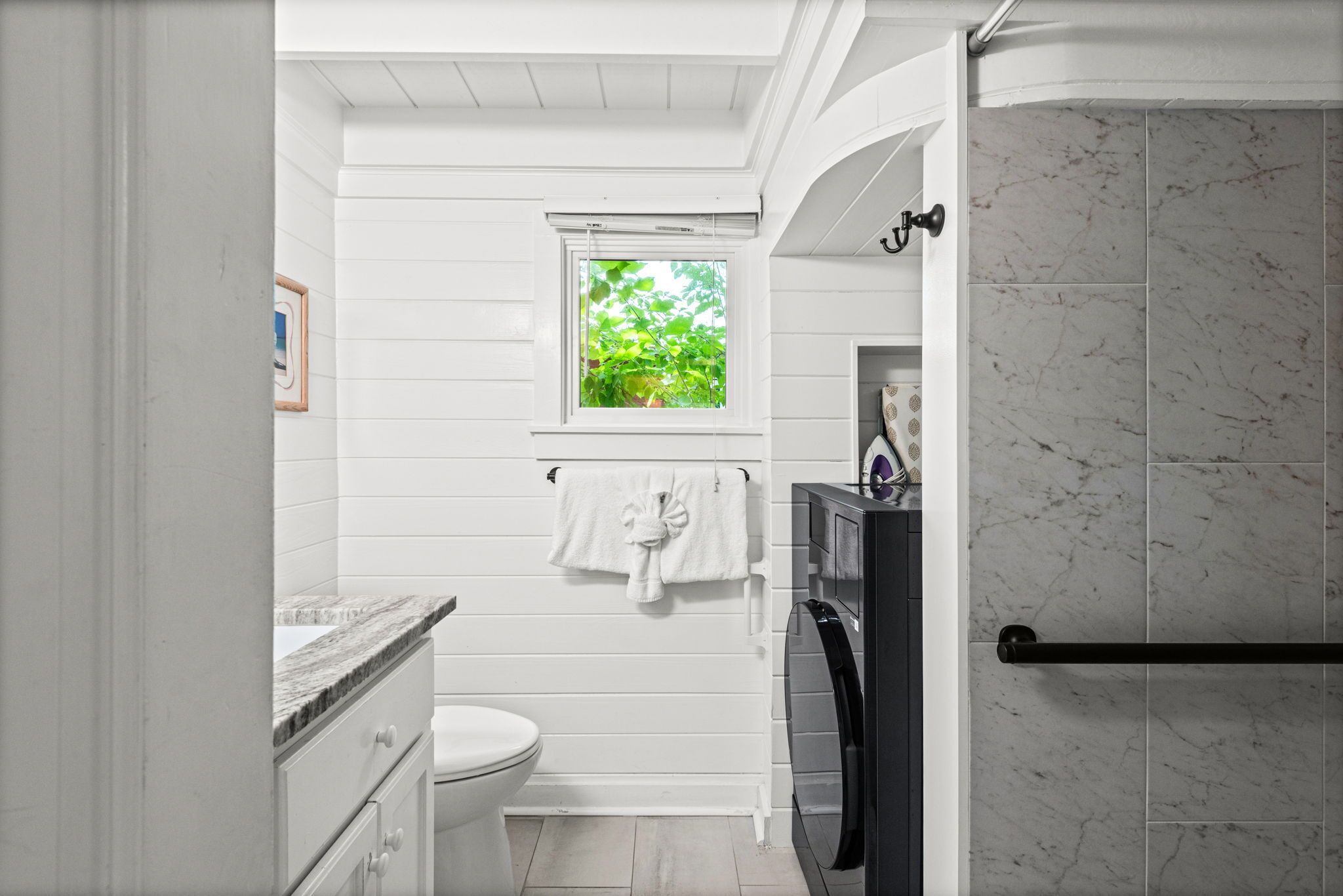 Ein Badezimmer mit Toilette, einem Fenster mit Blick auf Grünflächen, einem Handtuchhalter und einer Duschkabine mit Marmorfliesen.