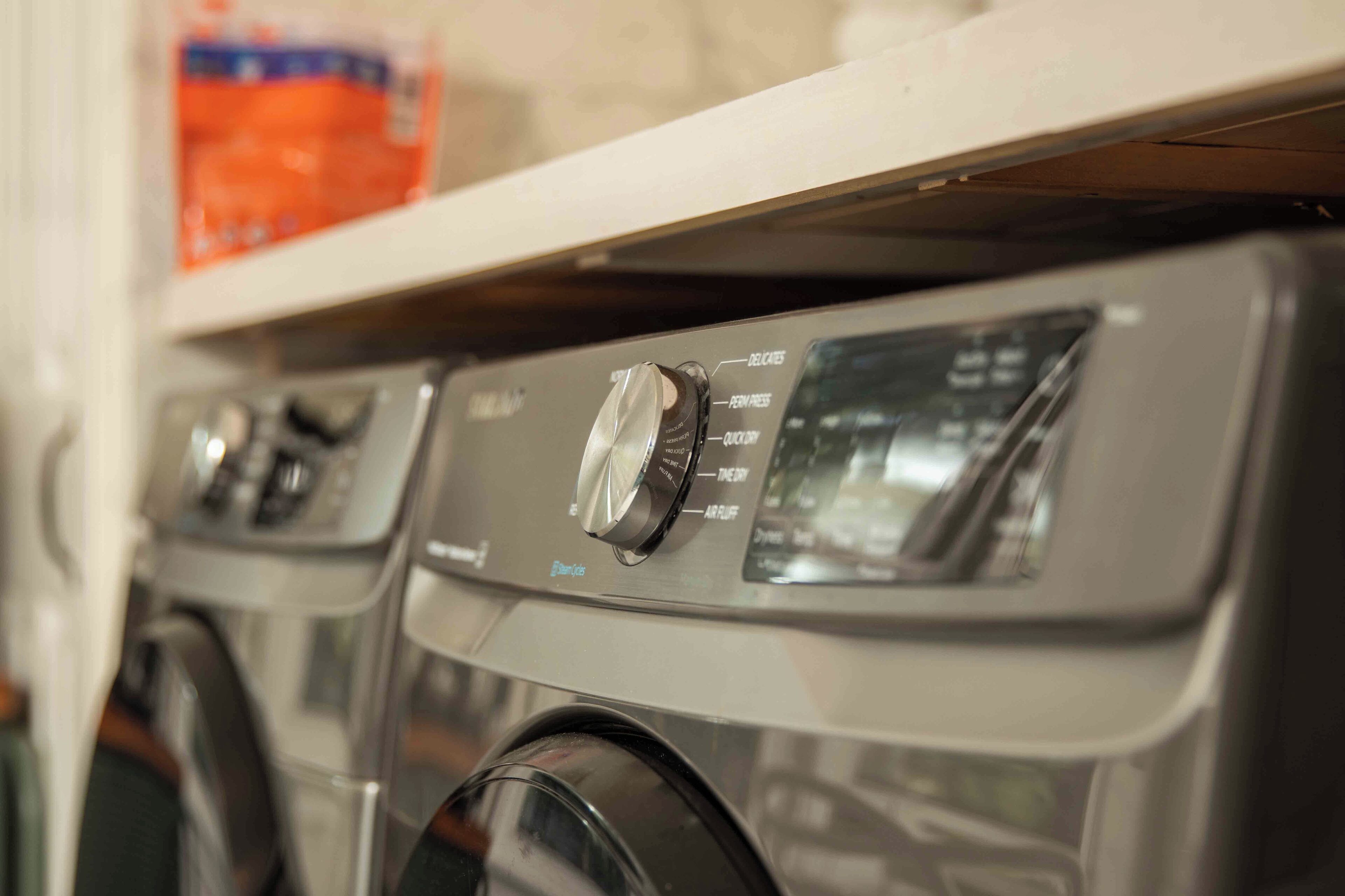 A row of front-loading washing machines with control panels and a shelf above them.
