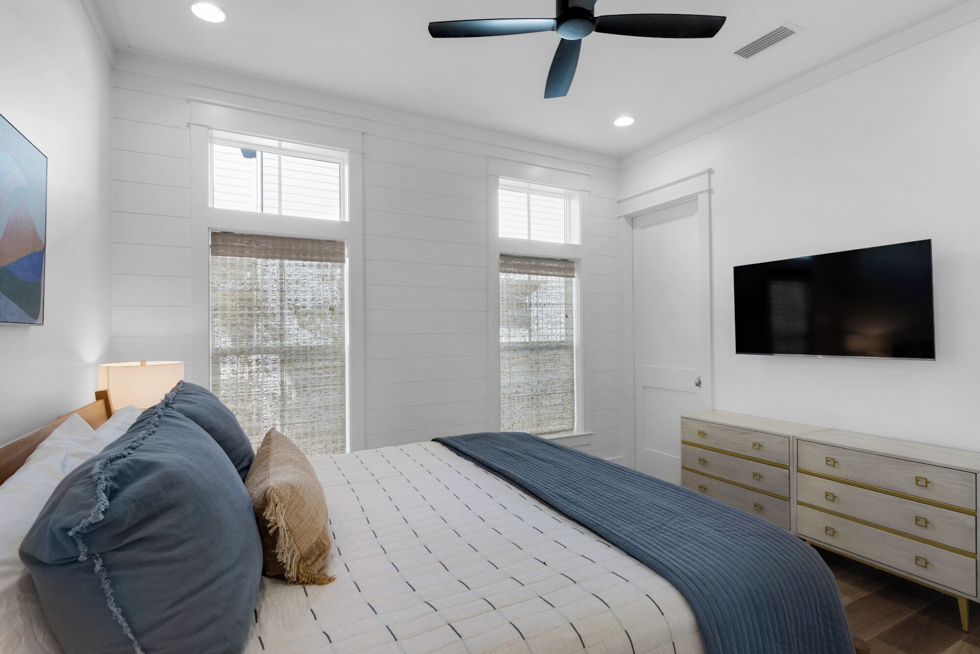A bedroom with a bed, a TV, a ceiling fan, and large windows.