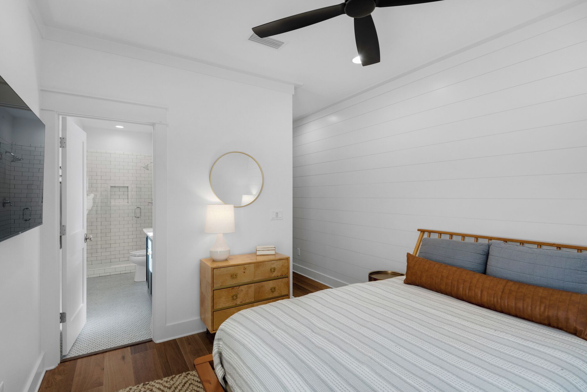 A bedroom with a bed, a wooden nightstand, a round mirror, and a ceiling fan.