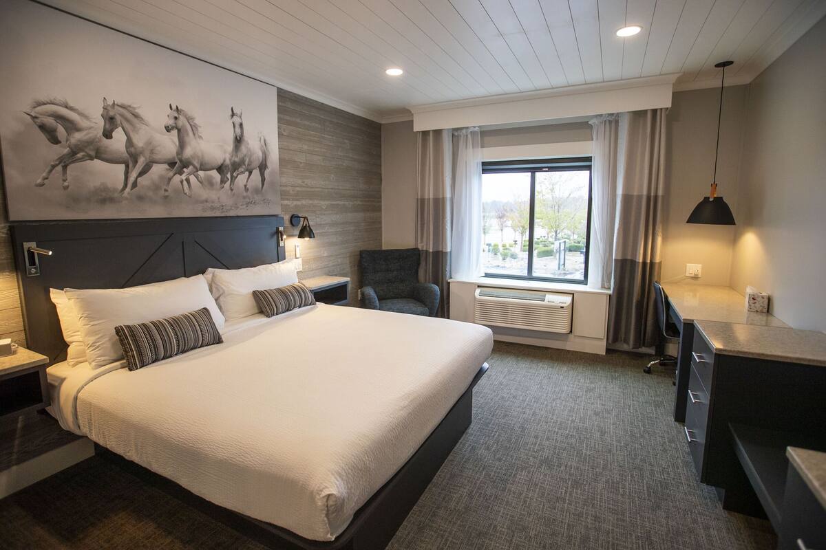 A modern hotel room with a large bed, a desk, a chair, and a painting of horses on the wall.