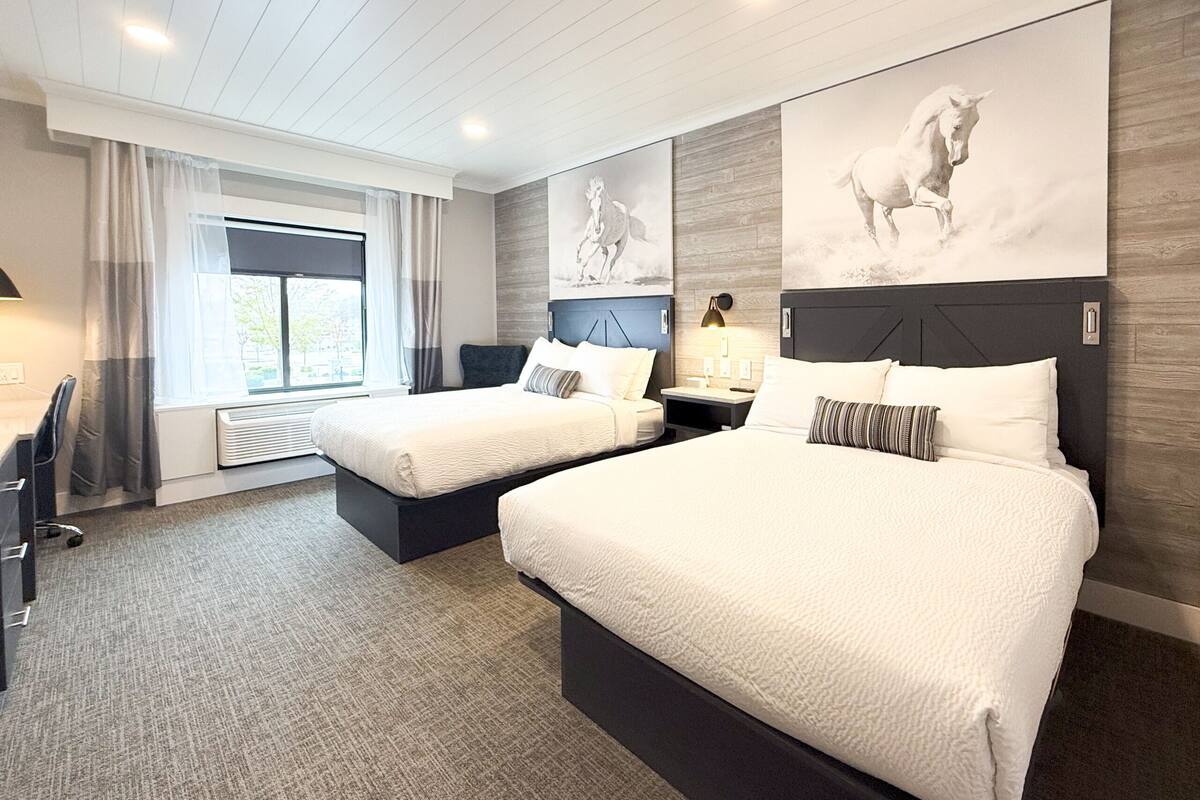 A hotel room with two beds, a desk, a chair, a window with curtains, and artwork on the walls.