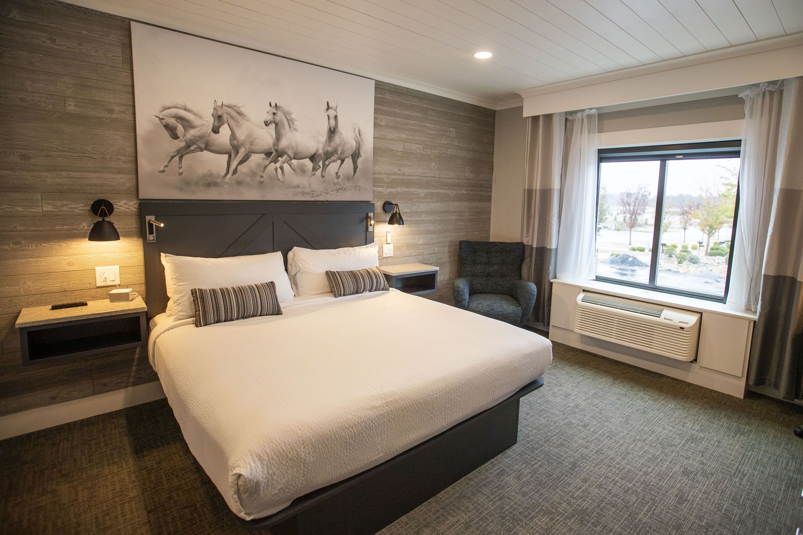 A bedroom with a large bed, a headboard with a horse painting, two bedside lamps, a chair, a window with curtains, and a view of the outdoors.
