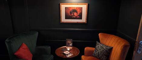 Ein dunkler Raum mit zwei Stühlen, einem kleinen Tisch und einem gerahmten Bild an der Wand.