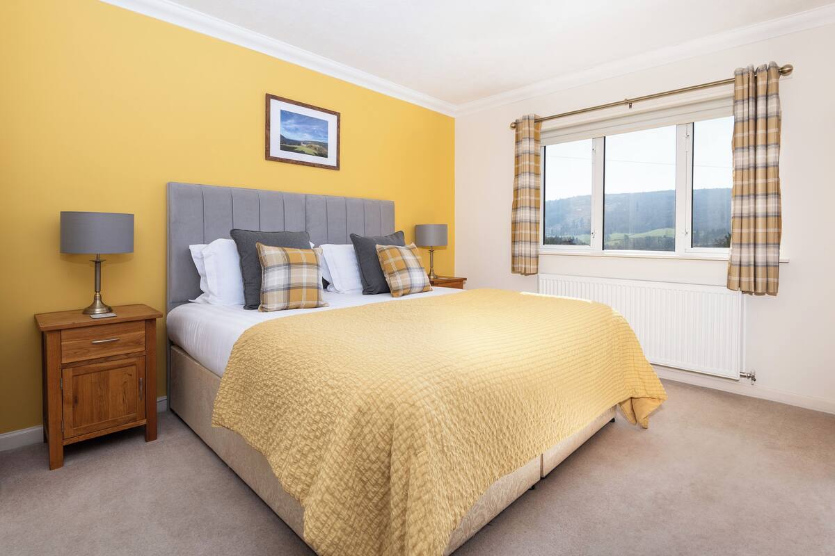 Quarto com cama, mesas de cabeceira, janela com cortinas e um quadro na parede.