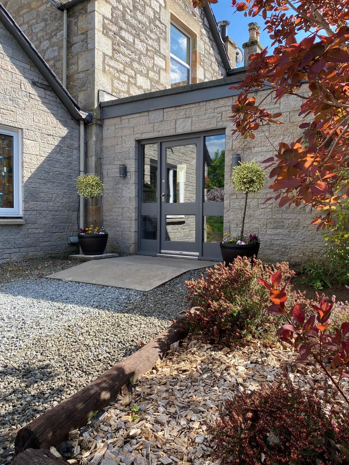A stone building with a modern entrance, potted plants, and a gravel pathway.