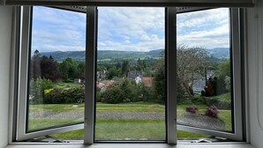 A room with a large window offering a view of a green landscape with trees and a distant hill. - Beinn Bhracaigh (Pitlochry)