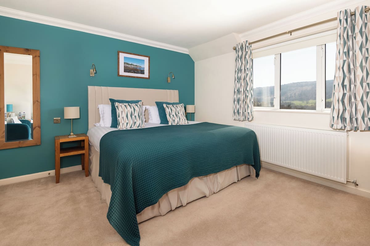 Quarto com cama, mesa de cabeceira, janela com cortinas e um quadro na parede.