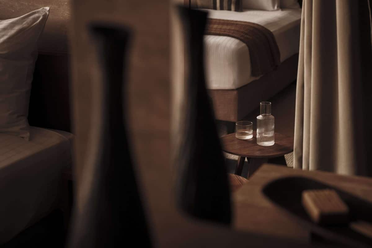 A modern hotel room with a bed, a bedside table with a bottle and glass, and a chair.