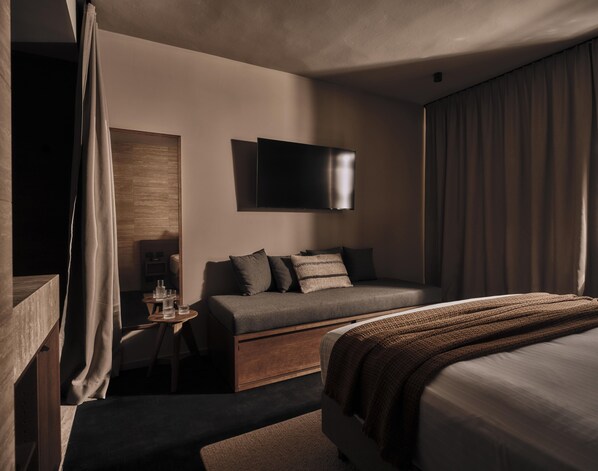 A modern hotel room with a sofa, a bed, a flat-screen TV mounted on the wall, and a small table with glasses. - Casa Cook Madonna - Adults Only (Pinzolo)