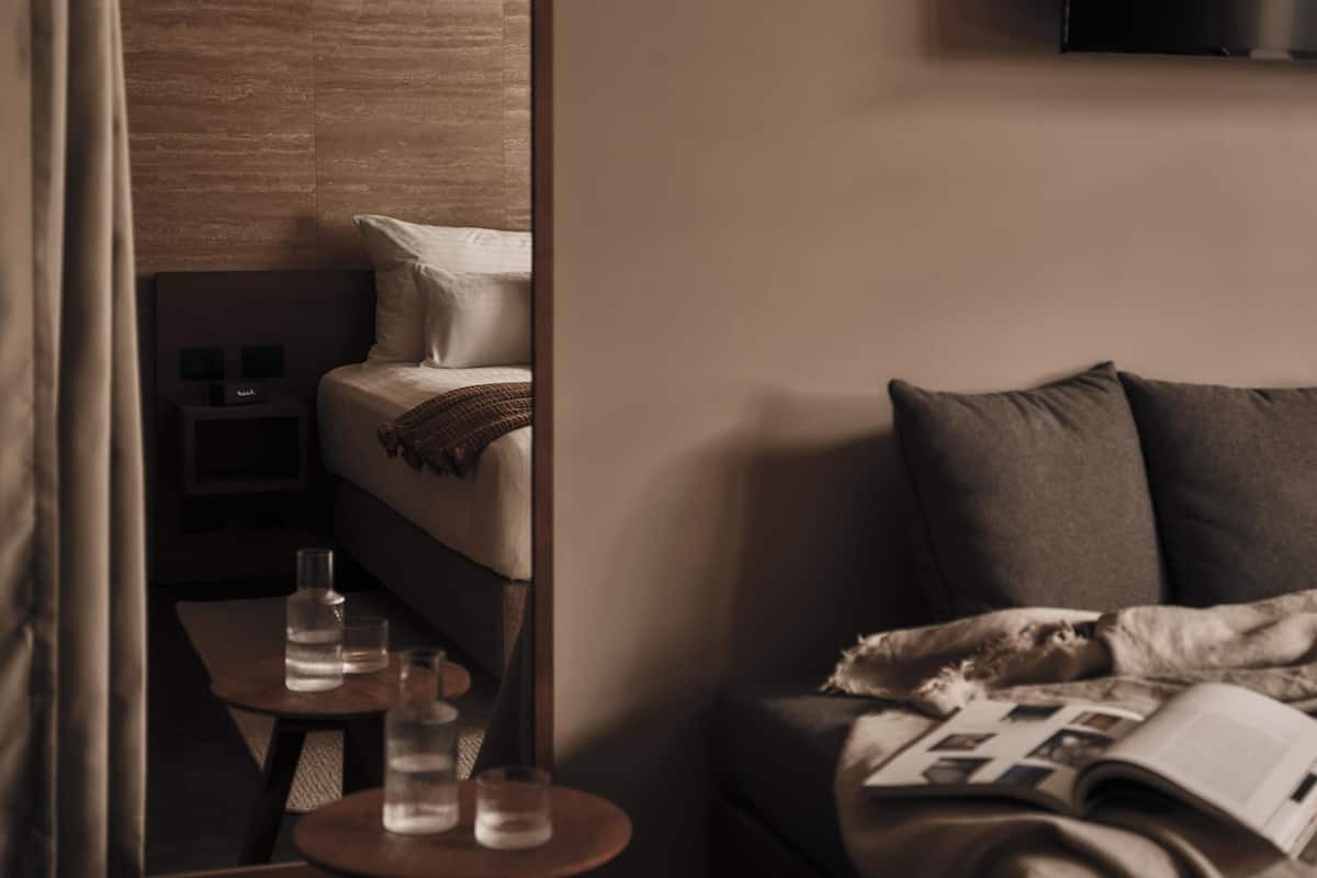 A hotel room with a large mirror, a bed with a blanket, a bedside table with glasses, and a TV on the wall.