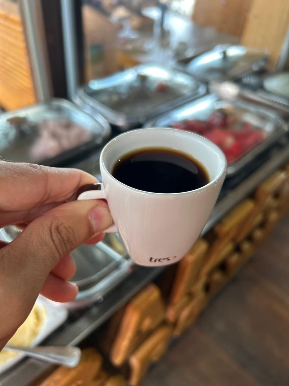 A hand holding a white mug with the word "tres" on it, filled with a dark liquid, likely coffee, in a setting with a buffet counter in the background.