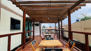 Eine hölzerne Außenterrasse mit Tischen und Stühlen, einer Pergola darüber und Blick auf die Skyline im Hintergrund.
