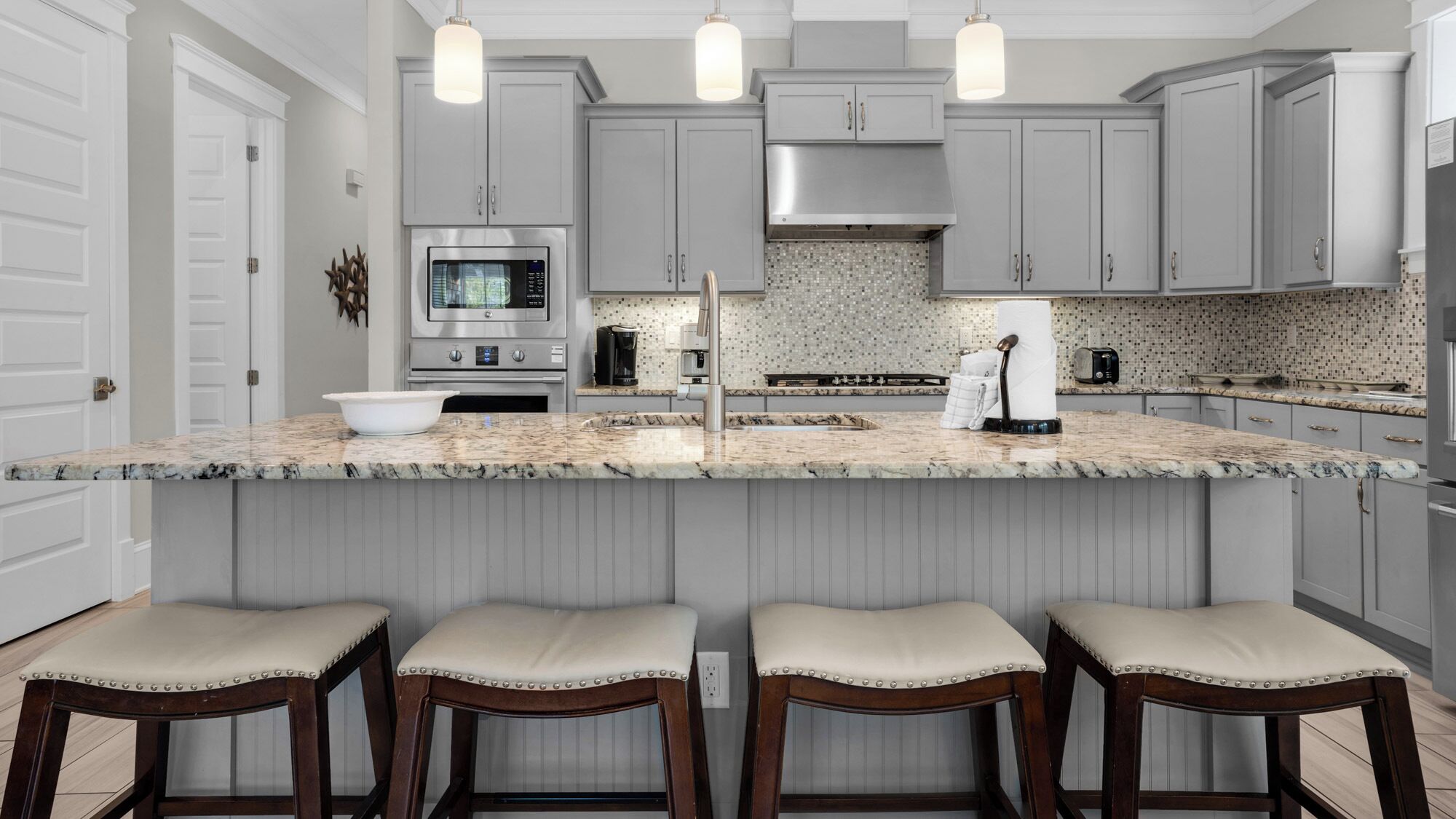 A modern kitchen with grey cabinets, a large island with stools, stainless steel appliances, and pendant lighting.