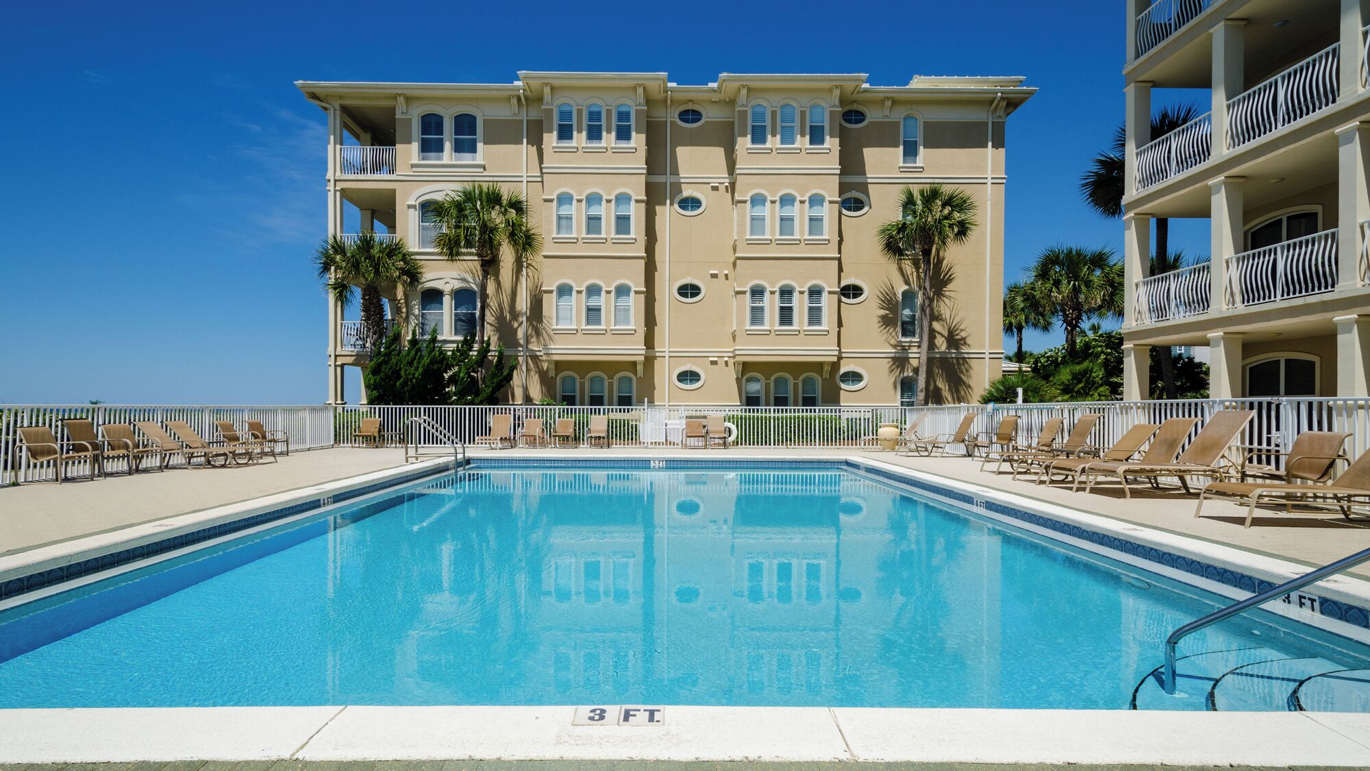 Piscina com marcação de profundidade de 3 pés, cercada por espreguiçadeiras e um edifício com varandas.