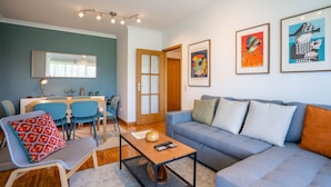 A modern living room with a grey sofa, a wooden coffee table, and a dining area with blue chairs and a wooden table. - Infante Cozy 1BR w/ Parking by LovelyStay (Vila Nova de Gaia)