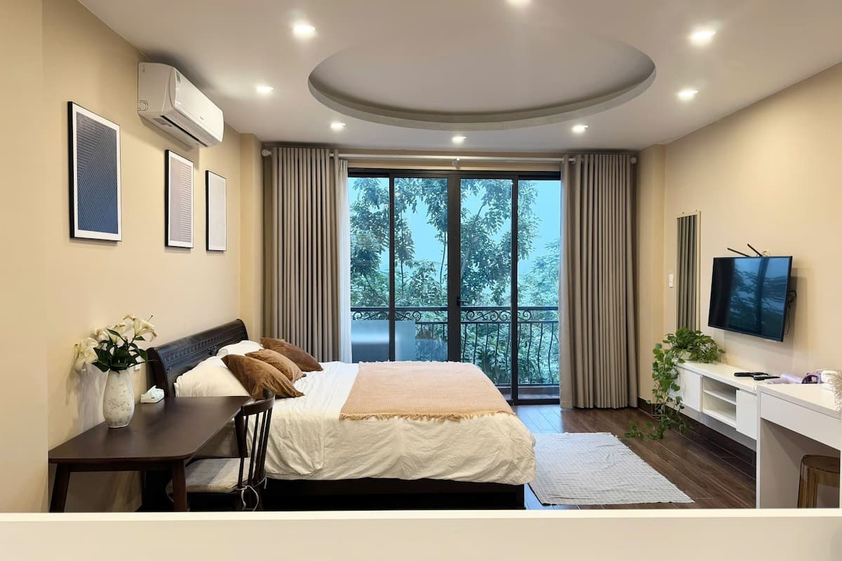 A modern hotel room with a large bed, a desk, a TV, and a balcony with a view of trees.