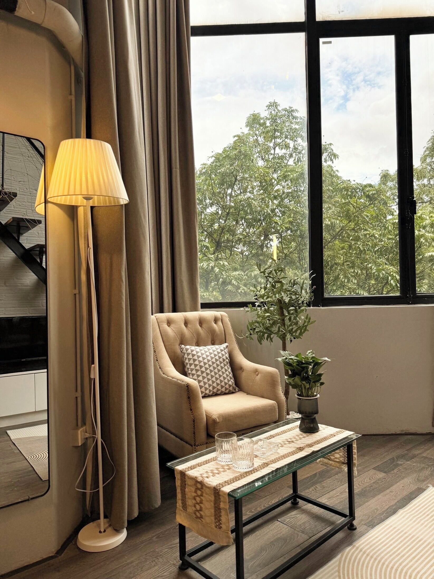 Une pièce comprenant un canapé, une lampe, une table et offrant une vue sur les arbres à travers de grandes fenêtres.